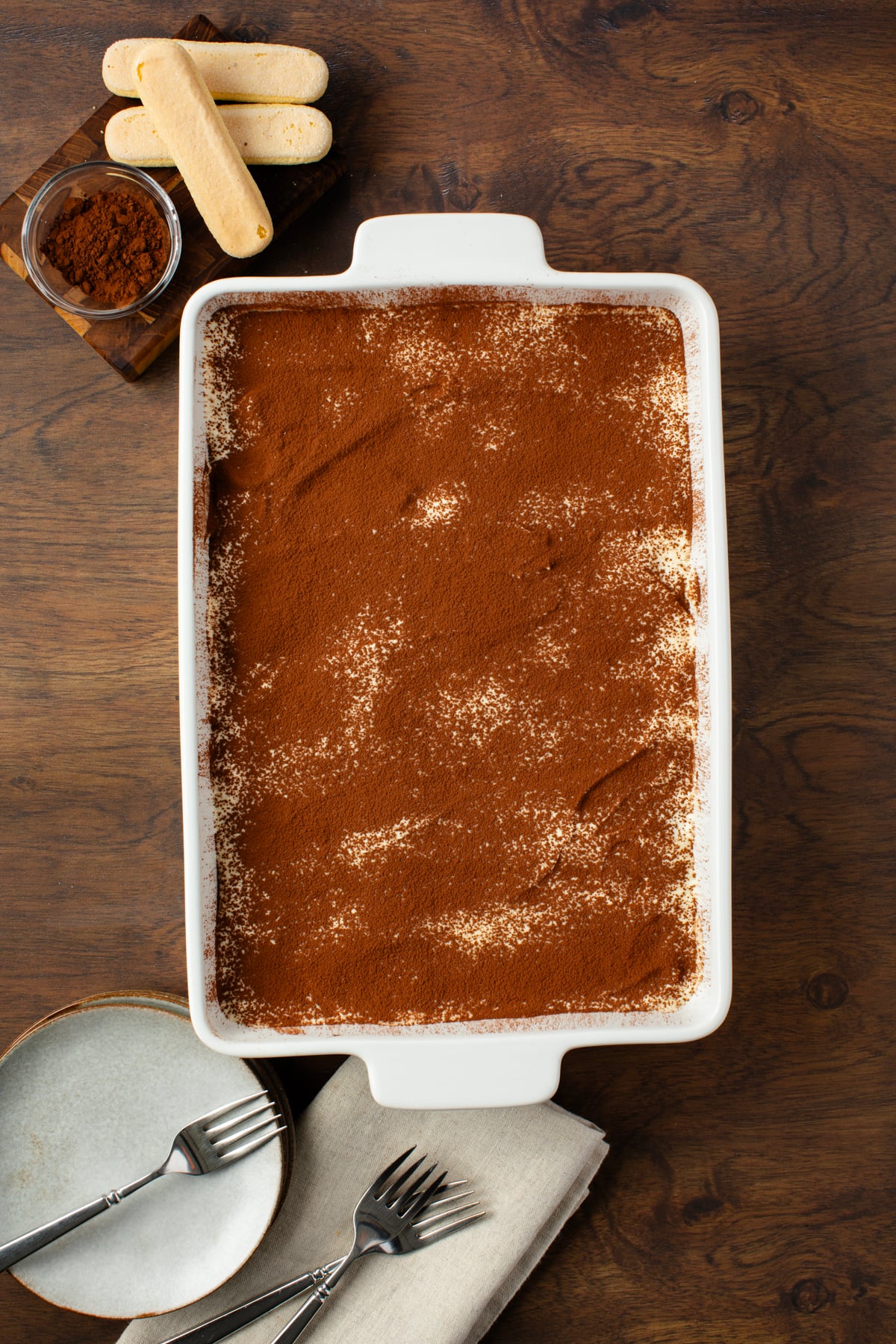 Your tiramisu is all done and a layer of chocolate powder is sprinkled over it. Dessert plates with forks are alongside the dish. 