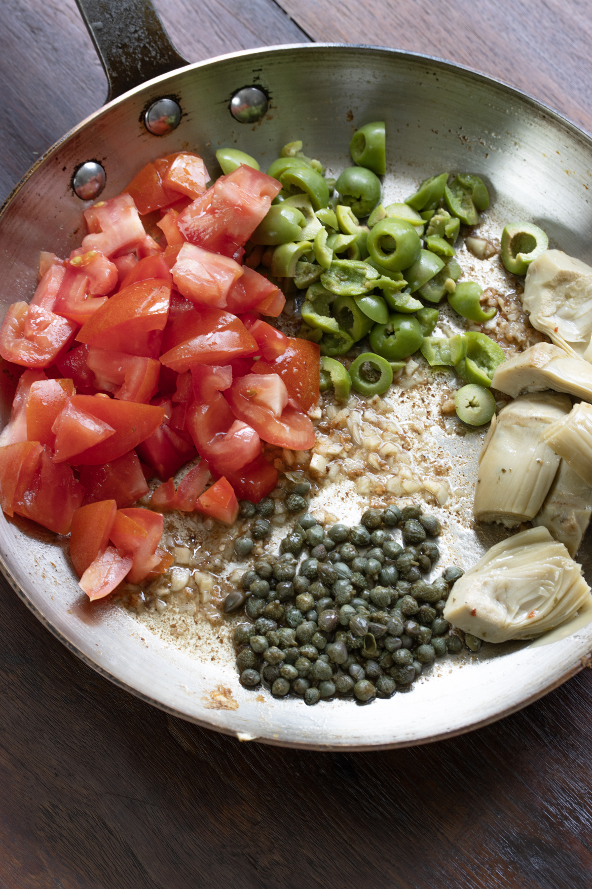 Cut tomatoes, olives, capers and artichokes hearts are placed in the skillet with the garlic. 