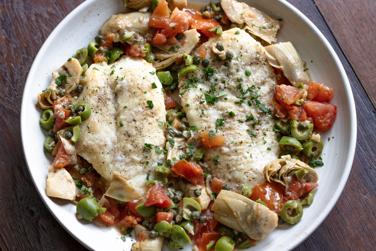 Cod piccata with Artichokes, tomoates and olives served in a round white serving plate. 