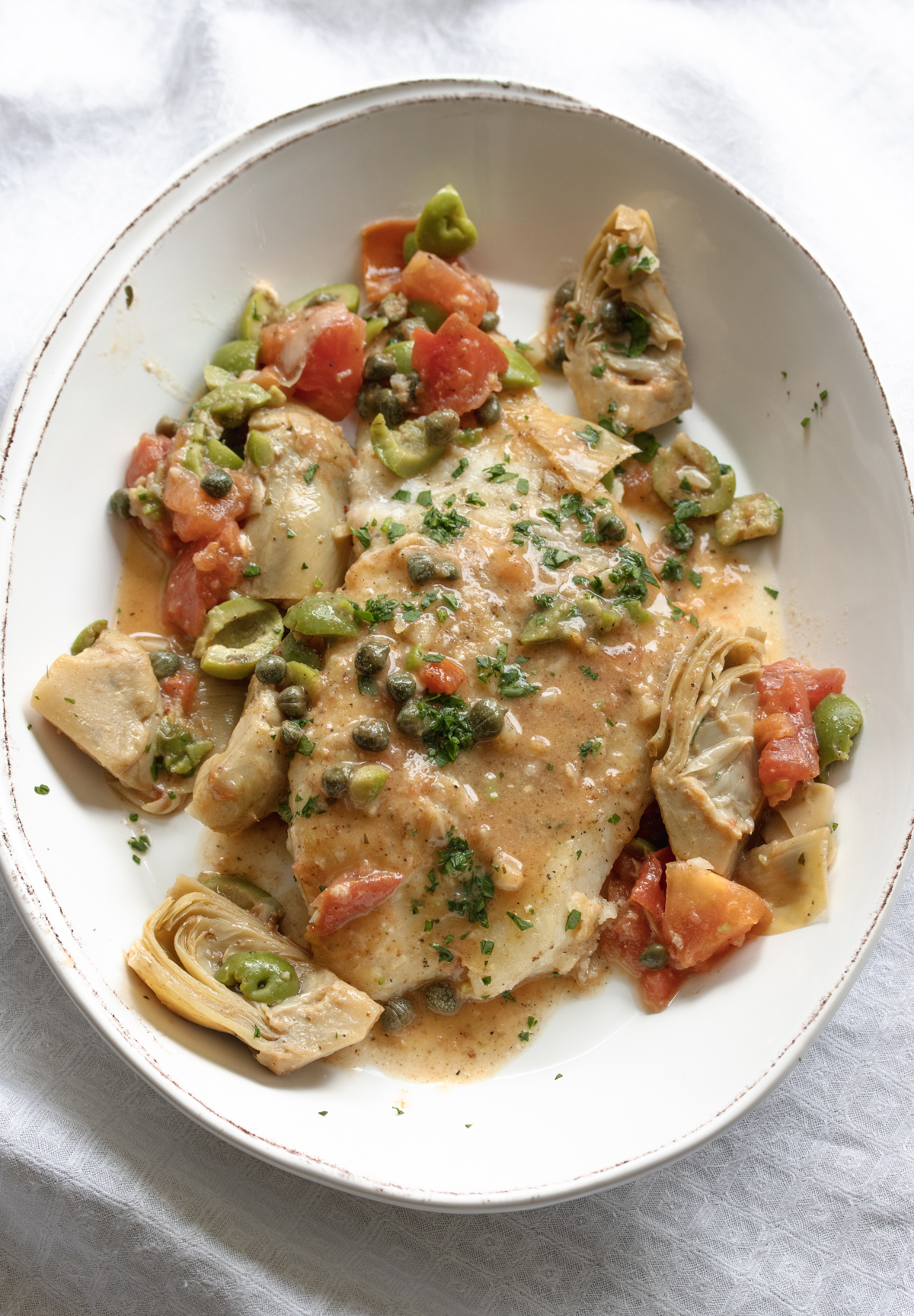 Single portion of cod piccata served in an oval dish with the artichokes, tomatoes, capers wine sauce over it and around it. 