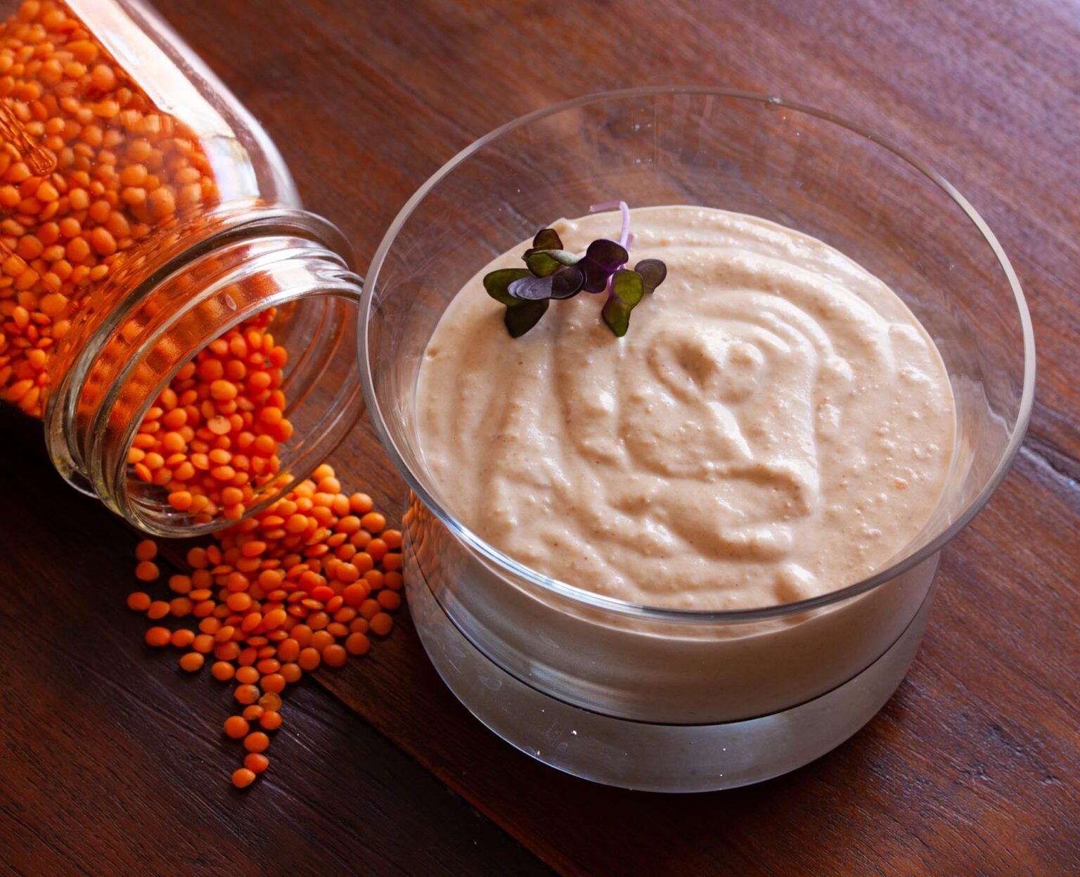 Red lentil hummus served in a glass small bowl. Jar of red lentils is next to it. 