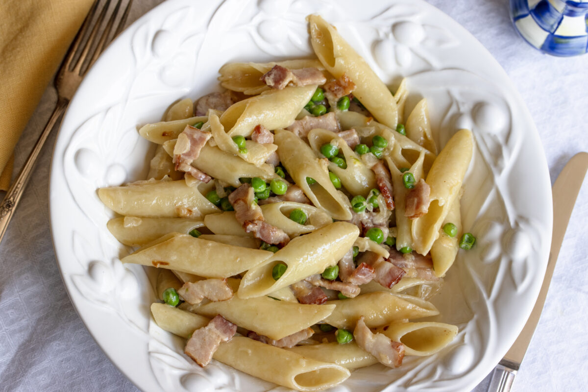 Penne pasta surrounded by pease and bacon and a cream sauce served on a white pasta plate.