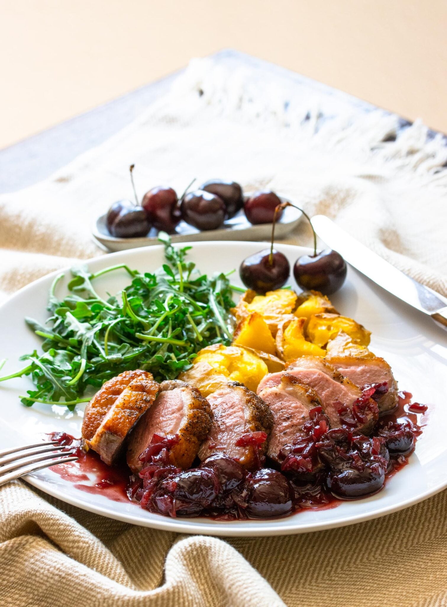 Duck breast with port and cherry sauce served in a white plate aliongside arugula salad and smashed potatoes