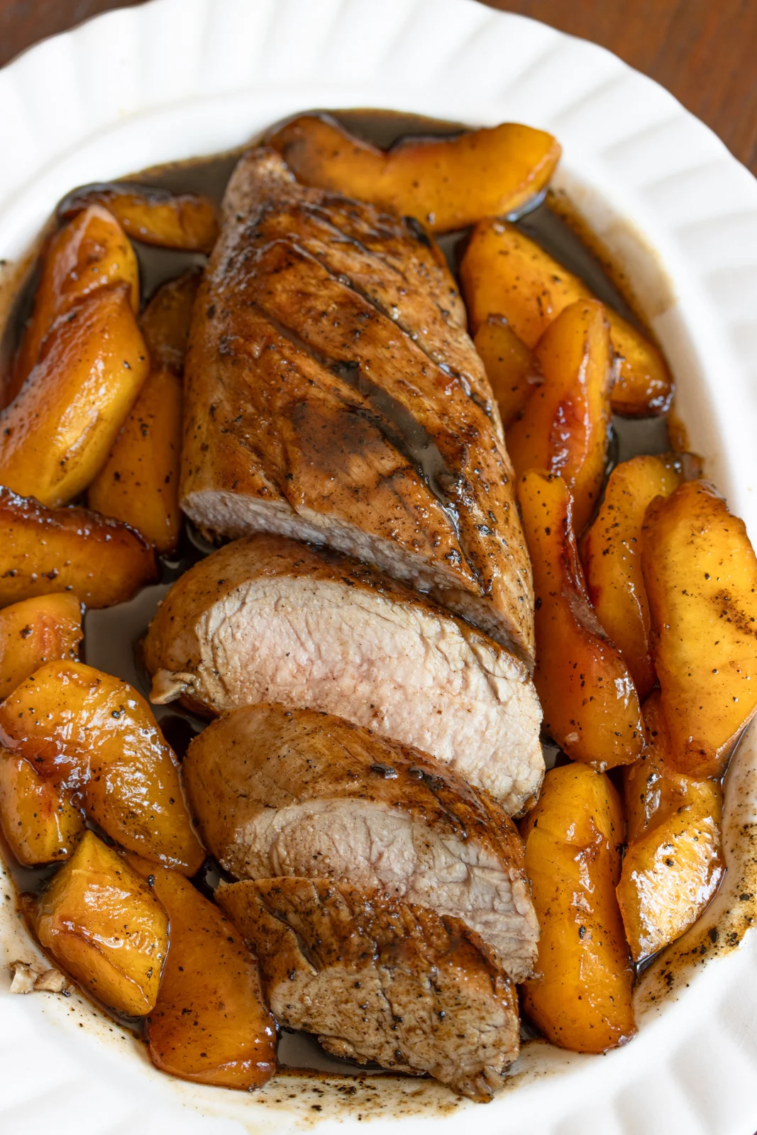 Glazed pork tenderloin half sliced and surrounded by peaches on a white serving dish.