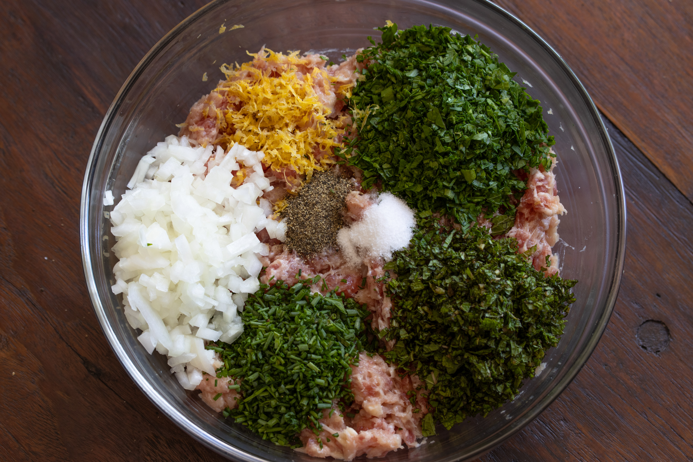 All the chopped herbs, lemon zest and onion are placed with the meat bowl to be blended. 