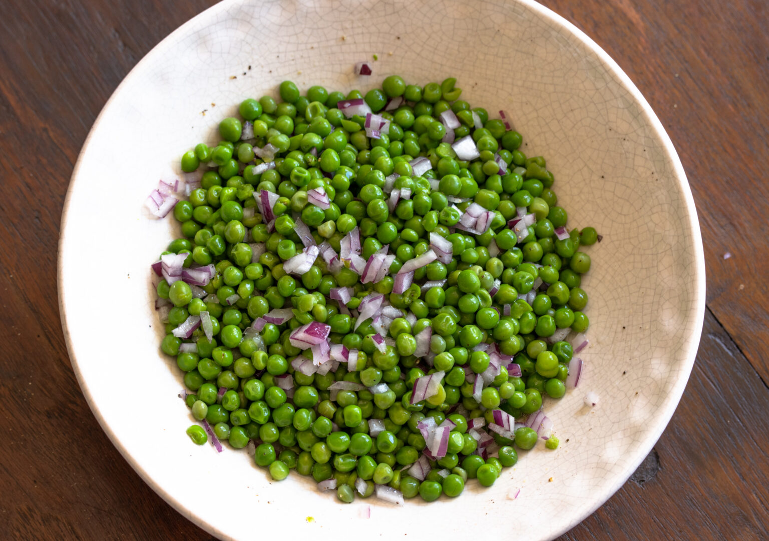 Olive oil, salt, pepper and red onions added to the cooked peas in a off white serving bowl.