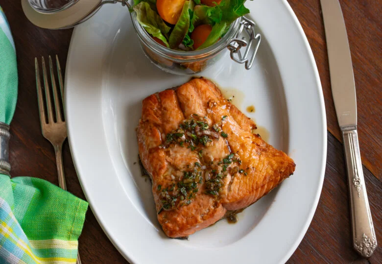 Broiled salmon with tarragon sauce over it and ready to be enjoyed. Silverware and napkin next to it.