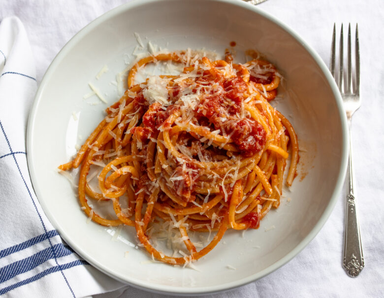 Amatriciana done and served in a white pasta bowl oever a white and blue tablecloth.