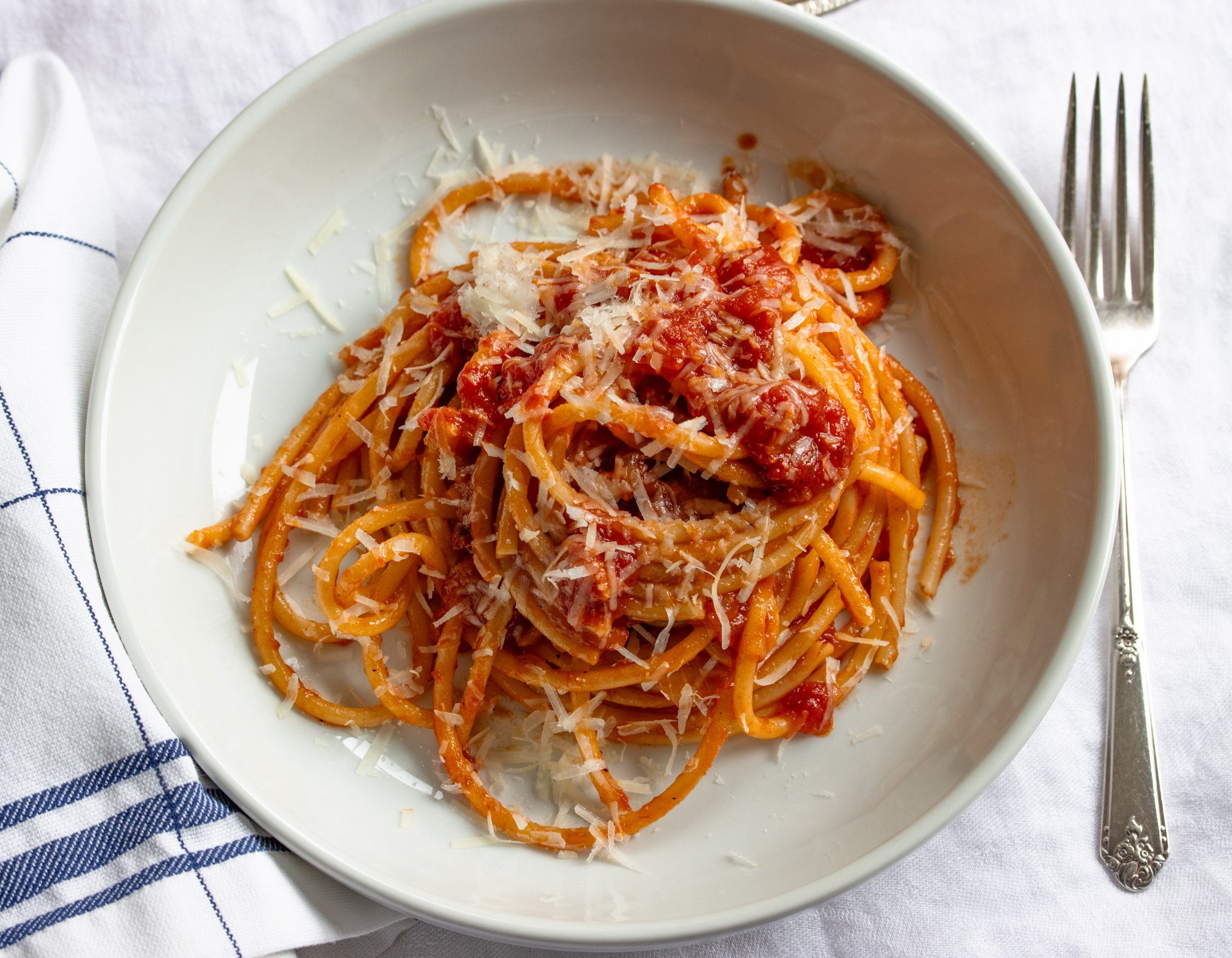 Amatriciana done and served in a white pasta bowl oever a white and blue tablecloth.