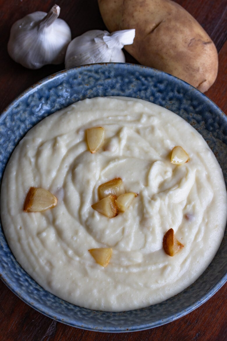 Garlic mashed potatoes served in a blue terracotta serving bowl surrounded by garlic and potatoes. 