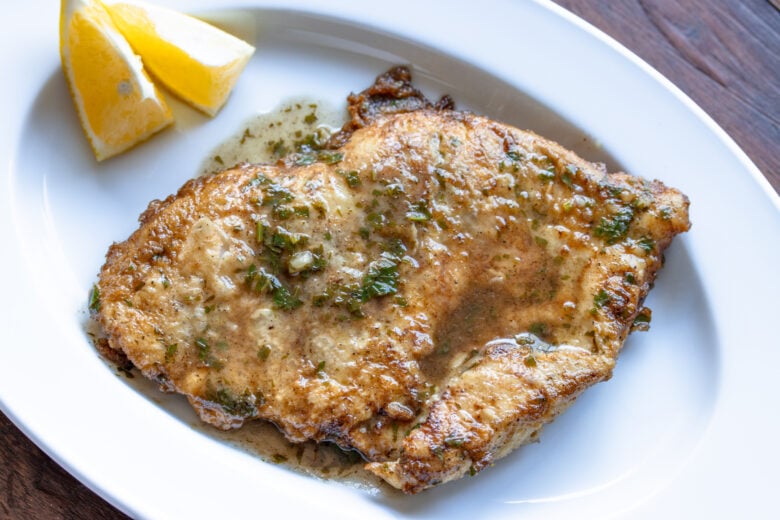 Chicken Francese served in a white oval dish with lemon wedges alongside and sauce poured over it.