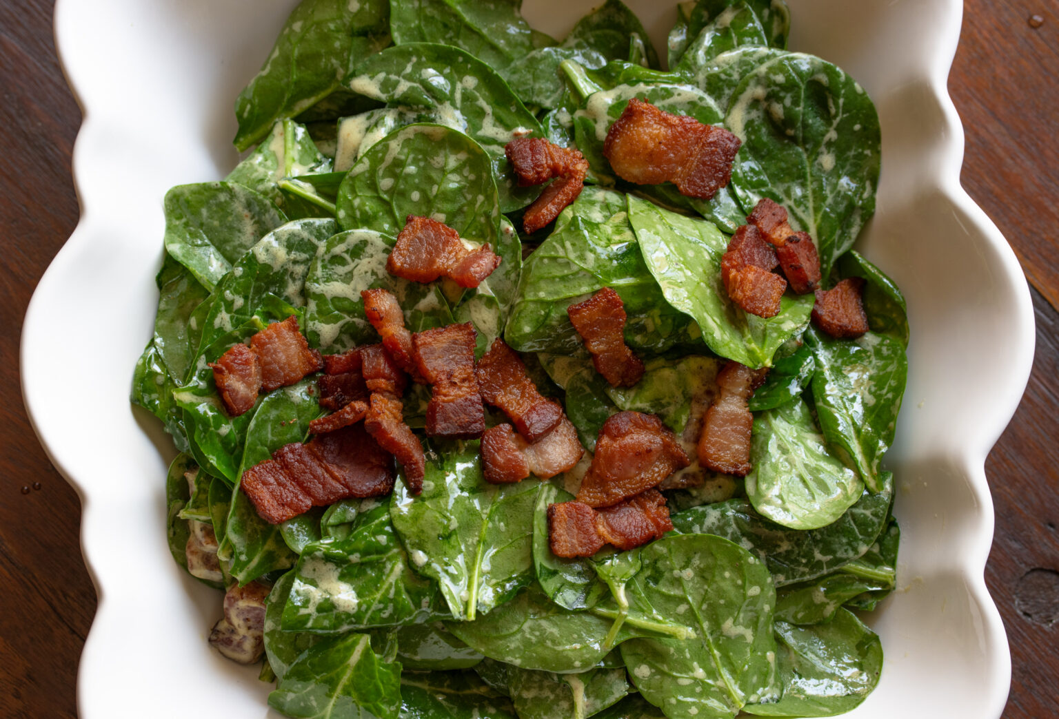 Spinach salad with warm bacon dressing served in a white scalloped serving bowl.