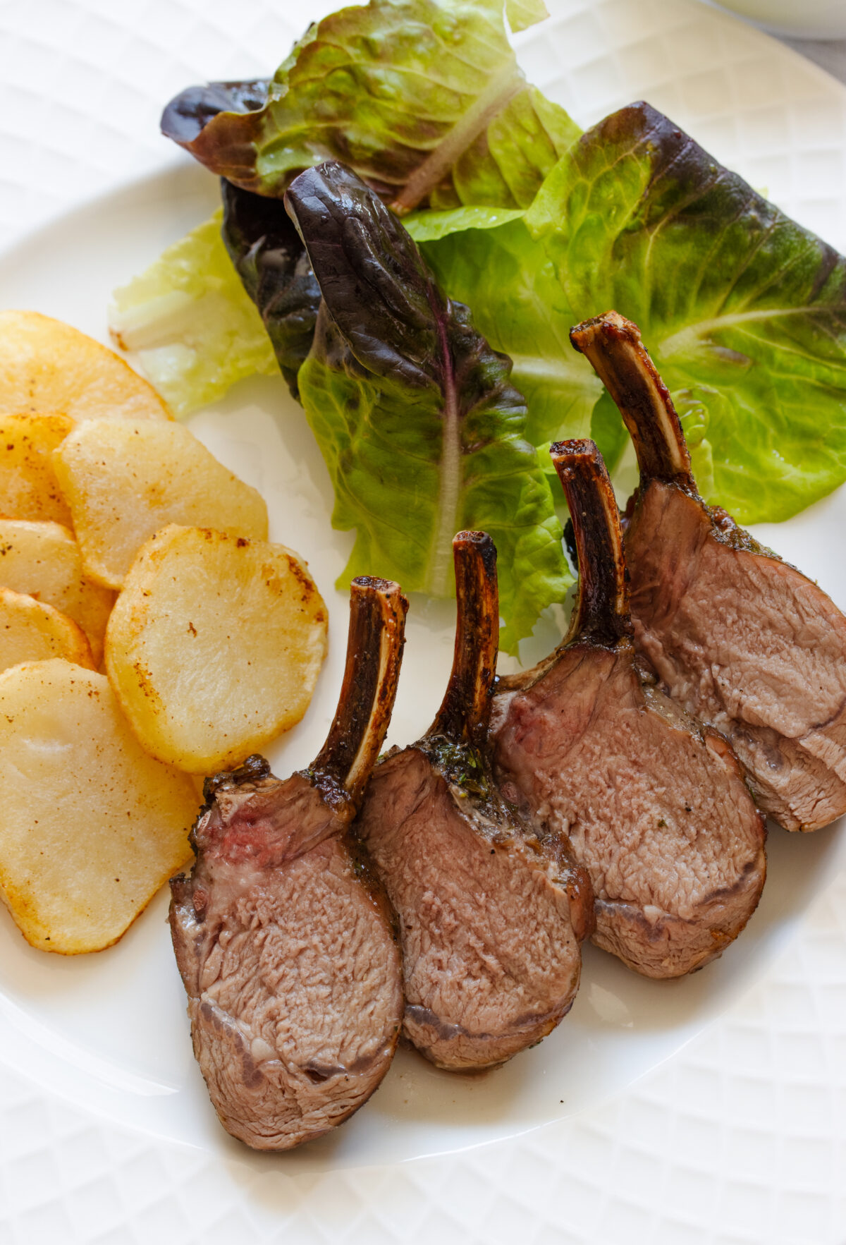 Roasted rack of lamb served alongside salad and potatoes on a white dish.