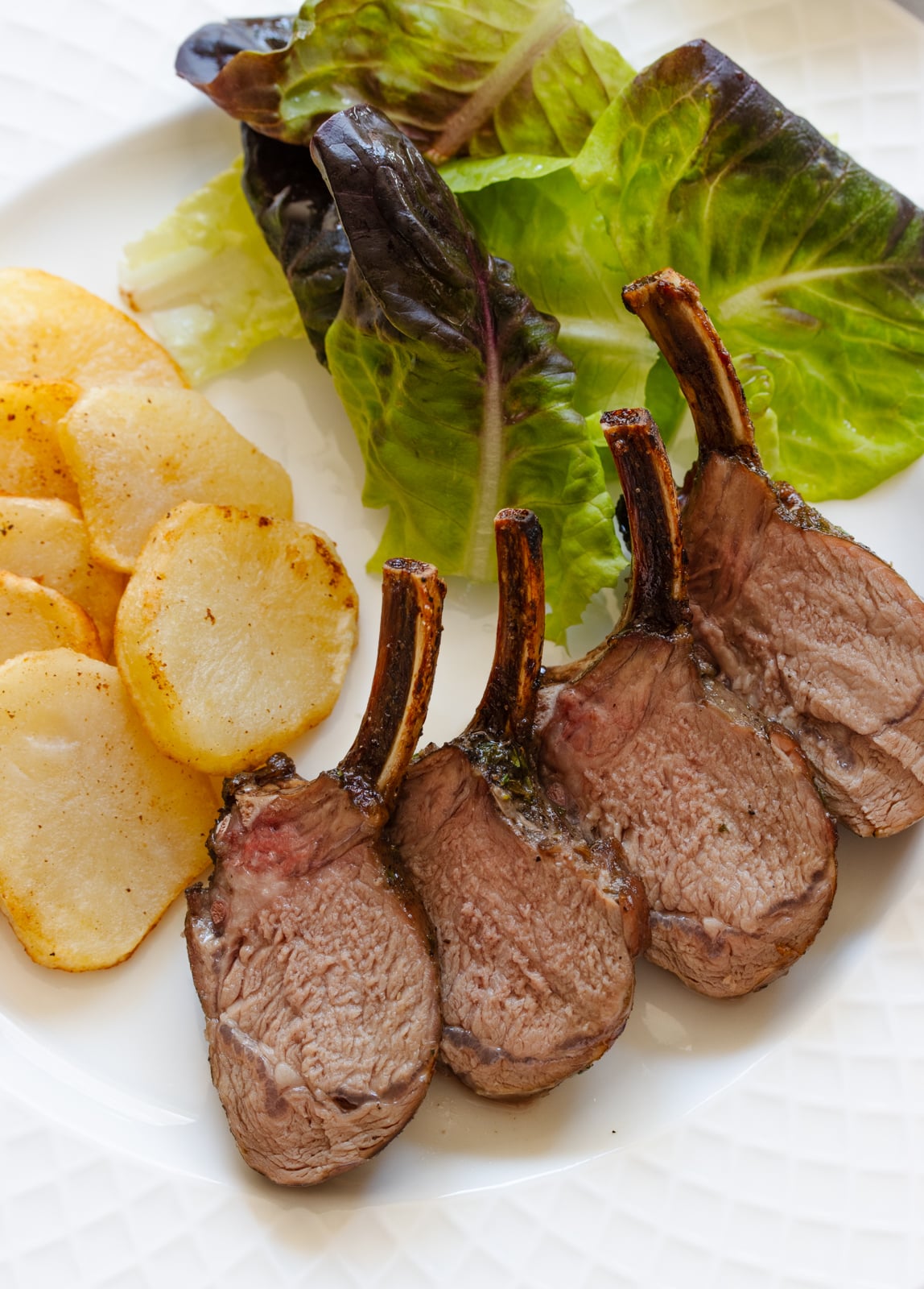 Roasted rack of lamb served alongside salad and potatoes on a white dish.
